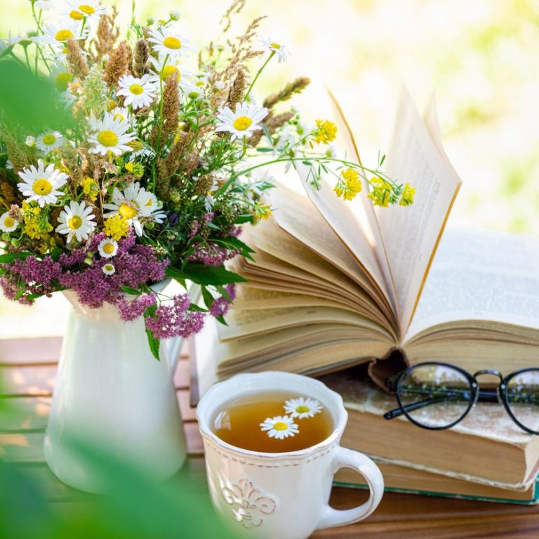 Tasse Tee, offene Buchseite, Blumenstrauß und Brille auf einem Tisch im Freien.