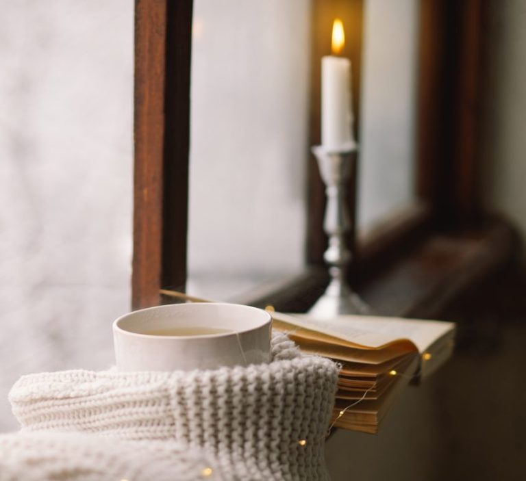 Eine Tasse Tee, eine brennende weiße Kerze, ein aufgeschlagenes Buch und eine Decke liegen auf einem Fensterbrett, gemütliche Atmosphäre.