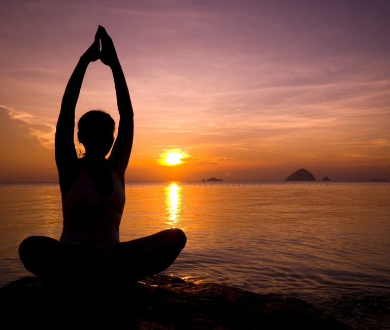 Silhouette einer Person in meditativer Pose bei Sonnenuntergang am Wasser.