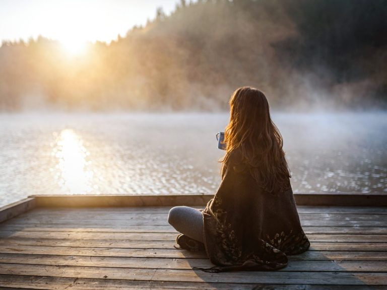 Frau sitzt am Ufer eines Sees und genießt den Sonnenaufgang mit einer Tasse Tee in der Hand.