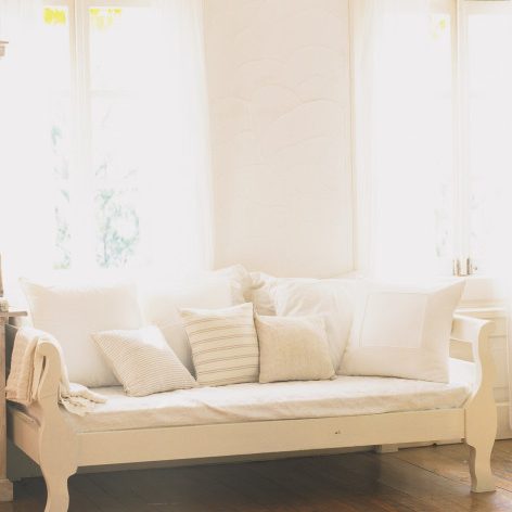 Helles Sofa in einem lichtdurchfluteten Zimmer mit vielen Kissen vor einem großen Fenster.